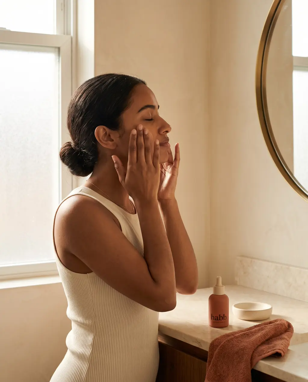 A woman applying skincare in soft morning light, glow visible on cheekbones.